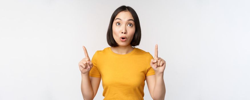 Happy beautiful asian girl showing advertisement, pointing fingers up, standing over white background.