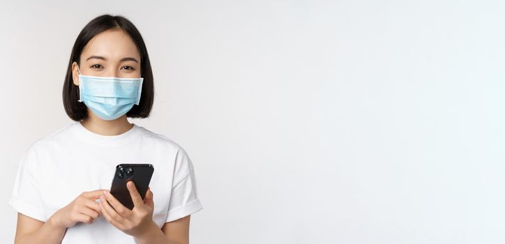 Health and covid-19 concept. Asian girl in medical face mask, using mobile phone application on quarantine, standing over white background.