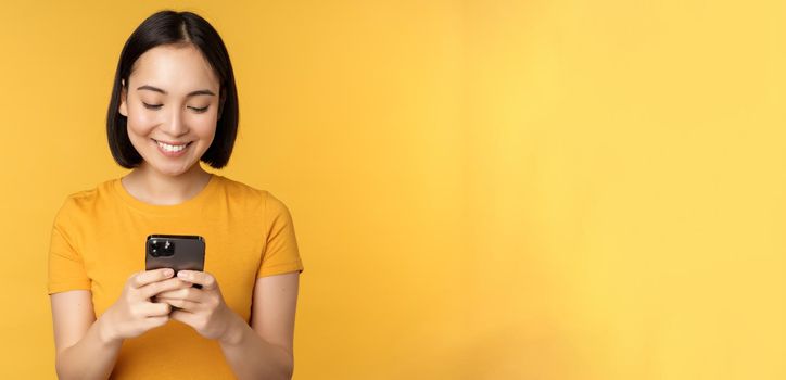 Technology. Smiling asian woman using mobile phone, holding smartphone in hands, standing in t-shirt against yellow background.