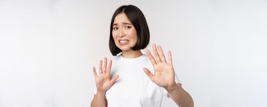 Disgusted asian woman rejecting smth, grimacing from dislike and aversion, stare with cringe, refusing offer, standing over white background.