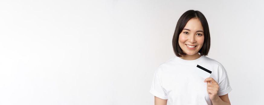 Portrait of korean smiling girl, bank client, showing credit card with happy face, standing against white background.