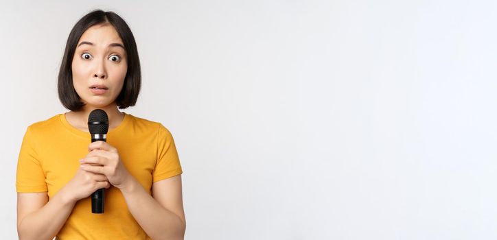 Modest asian girl holding microphone, scared talking in public, standing against white background. Copy space