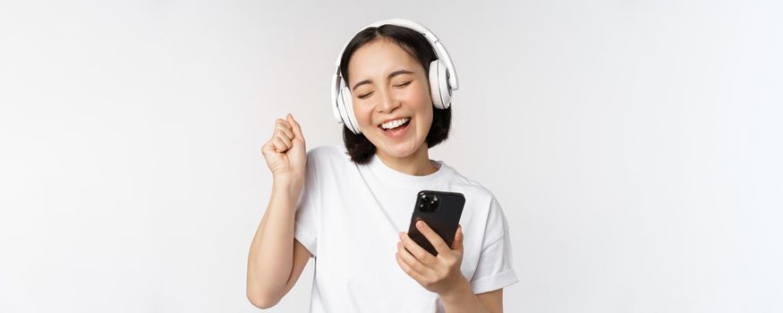 Beautiful modern asian girl, listening music in headphones, holding mobile phone, using smartphone app, standing over white background.