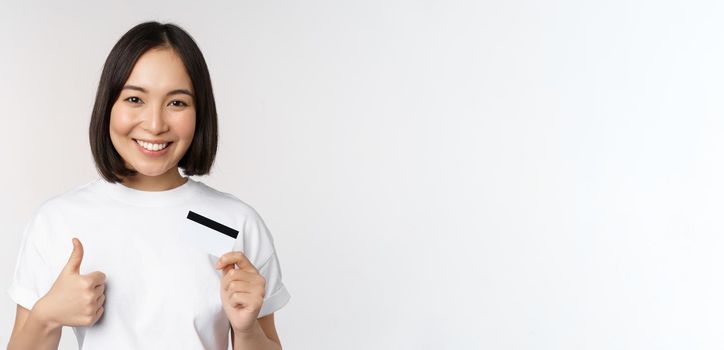 Portrait of beautiful young modern asian woman, showing credit card and thumbs up, recommending contactless payment, standing over white background.