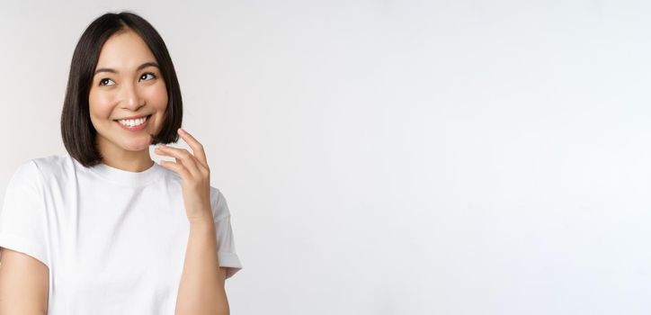 Portrait of cute coquettish woman laughing and smiling, looking aside thoughtful, thinking or imaging smth, standing in white t-shirt over studio background.