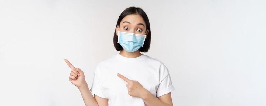 Surprised asian girl in medical mask, pointing fingers left, showing promo offer, raising eyebrows, amazed reaction, standing against white background.