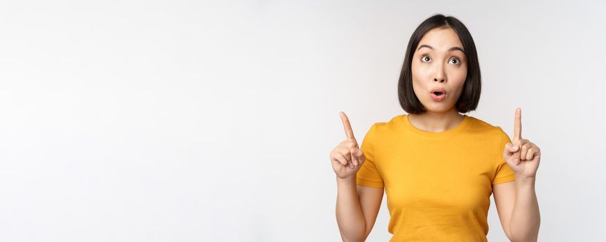 Happy beautiful asian girl showing advertisement, pointing fingers up, standing over white background.