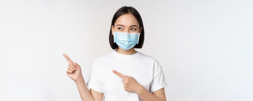 Young korean woman in medical face mask pointing fingers left and looking at logo, showing advertisement or banner, standing over white background.