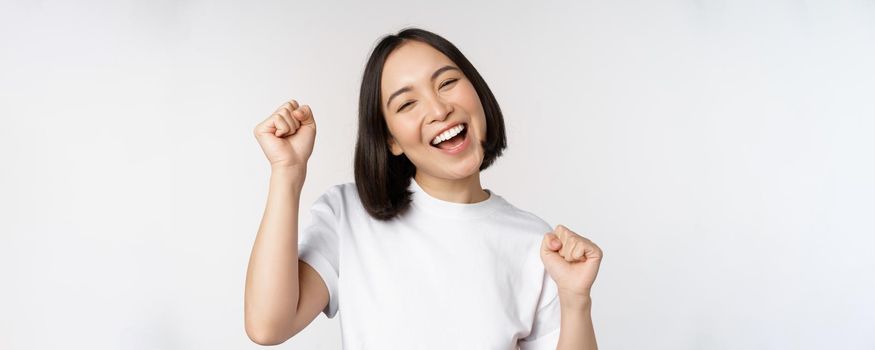 Dancing asian girl celebrating, feeling happy and upbeat, smiling broadly, standing over studio white background.