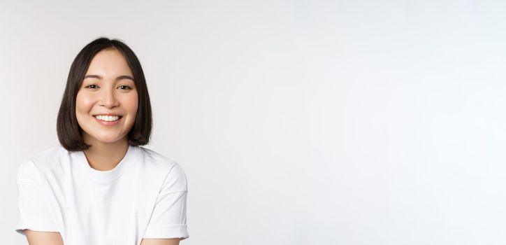 Beautiful korean girl smiling, white teeth, looking lovely at camera, standing in white tshirt over studio background.