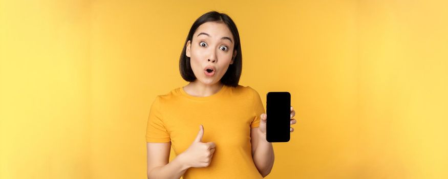 Excited asian woman showing mobile phone screen, thumbs up, like smth good, recommending smartphone application, standing over yellow background.