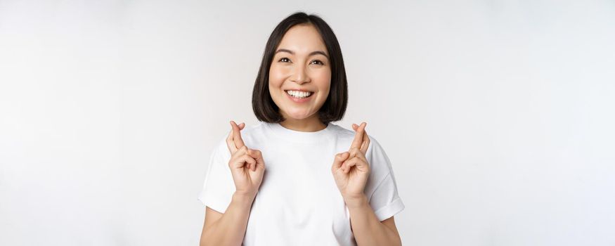 Hopeful asian girl cross fingers, making wish, anticipating, wishing smth, standing in tshirt over white background. Copy space