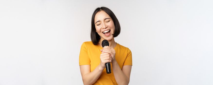 Happy asian girl singing and having fun, holding microphone at karaoke, standing in yellow tshirt against white background. Copy space