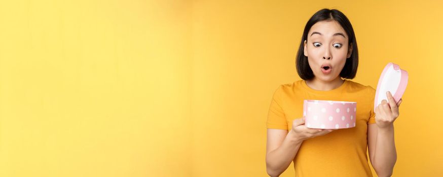 Portrait of excited asian woman, open gift box with surprised happy face, standing over yellow background.