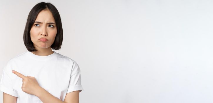 Angry and upset young asian woman pointing left, looking with regret or jealousy, feel unfair, standing over white background. Copy space