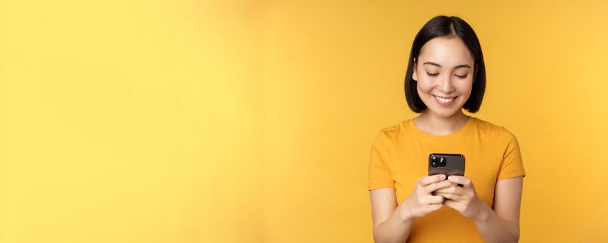 Technology. Smiling asian woman using mobile phone, holding smartphone in hands, standing in t-shirt against yellow background.