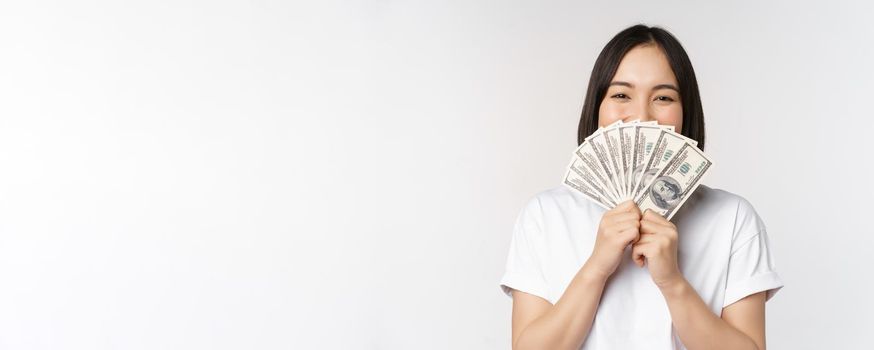 Portrait of smiling asian woman holding dollars money, concept of microcredit, finance and cash, standing over white background.