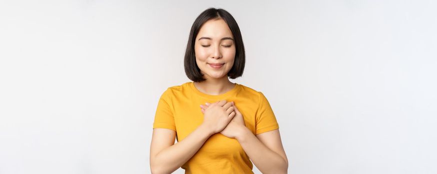 Romantic asian girfriend, holding hands on heart, smiling with care and tenderness, standing in yellow tshirt over white background.