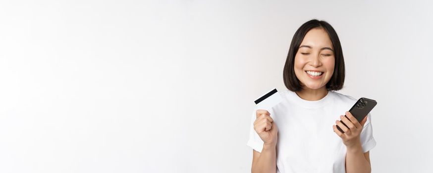 Online shopping. Happy asian woman using credit card and smartphone app, paying on website via mobile phone, white background.