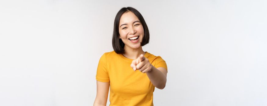 Happy beautiful asian woman laughing, pointing finger at camera and chuckle, smiling carefree, standing in yellow tshirt over white background.