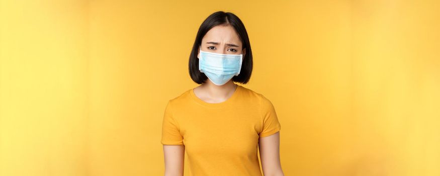 Image of sad and disappointed asian woman in medical mask, frowning and looking displeased, standing against yellow background.