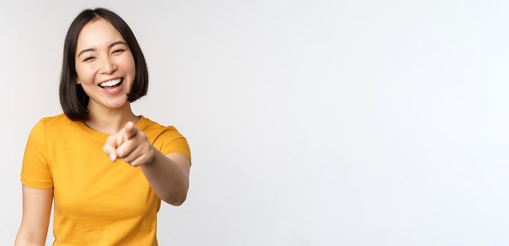 Happy beautiful asian woman laughing, pointing finger at camera and chuckle, smiling carefree, standing in yellow tshirt over white background.