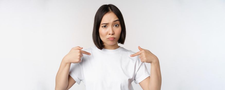 Young asian woman pointing at herself with disbelief, being chosen, surprised by her candidature, standing over white background.