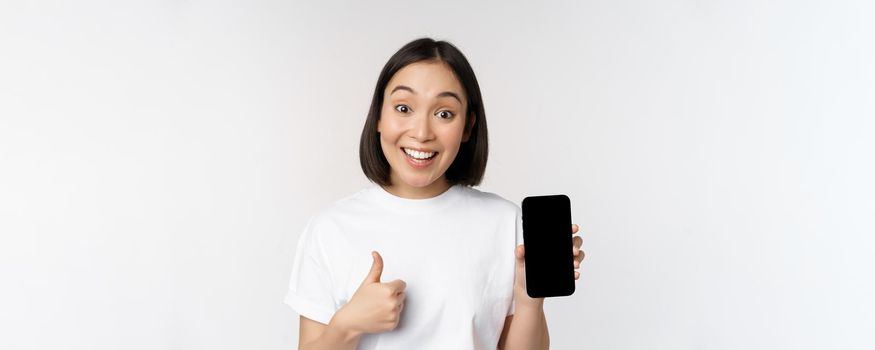 Enthusiastic young woman showing thumb up and mobile phone screen, standing in tshirt over white background. Copy space