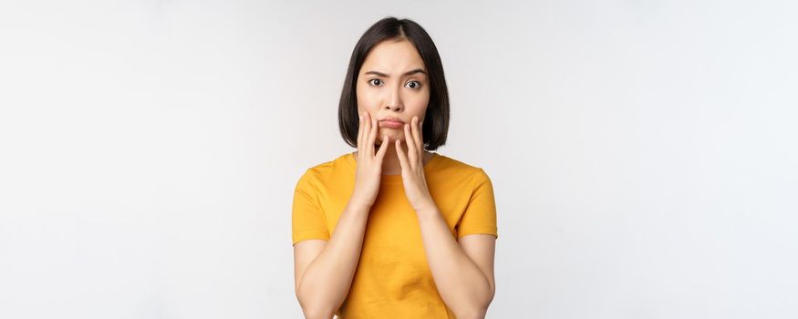 Shocked korean girl touching her face, looking concerned at camera, standing in yellow t-shirt over white background.