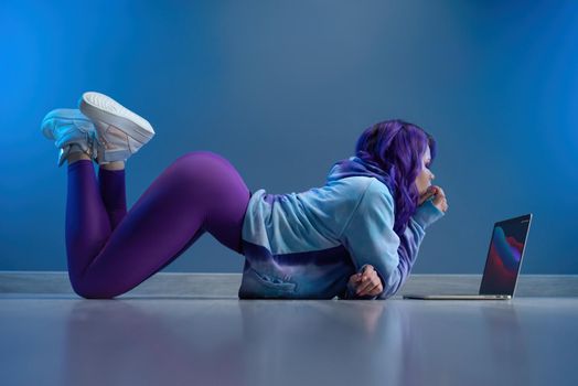 a girl in stylish purple sportswear and with purple hair is sexy lying on the floor with a laptop