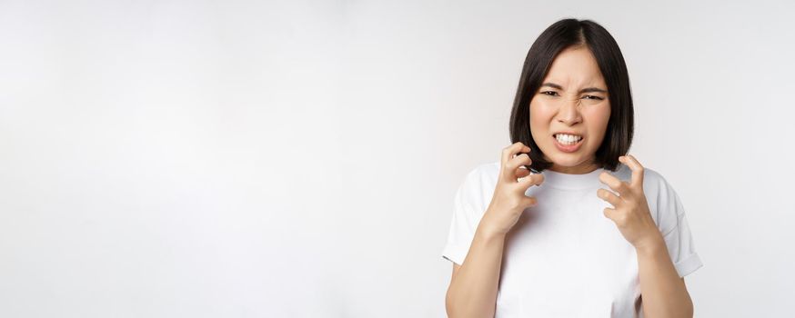 Angry asian woman cursing, looking outraged and annoyed, clench teeth and frowning furious, standing over white background.