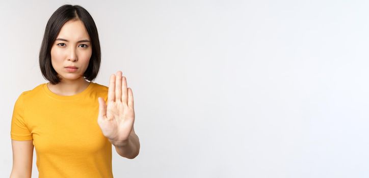 Portrait of asian woman looking serious and angry, showing stop prohibit gesture, taboo sign, forbidding smth, standing in yellow tshirt over white background.