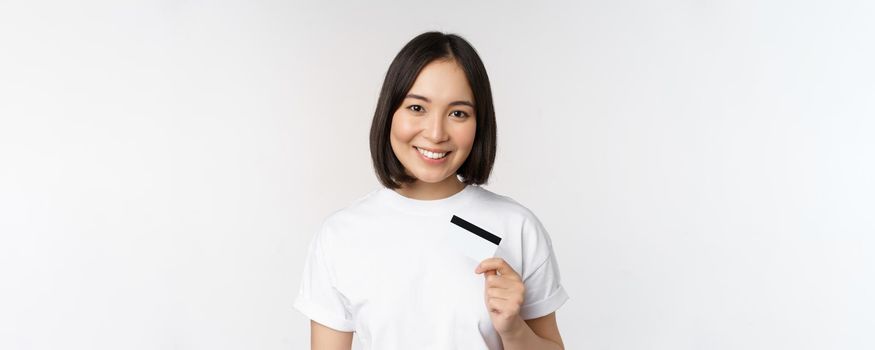 Portrait of korean smiling girl, bank client, showing credit card with happy face, standing against white background.