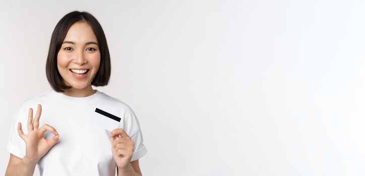 Smiling korean girl showing okay sign and recommending credit card of copy space bank, standing in tshirt over white background.