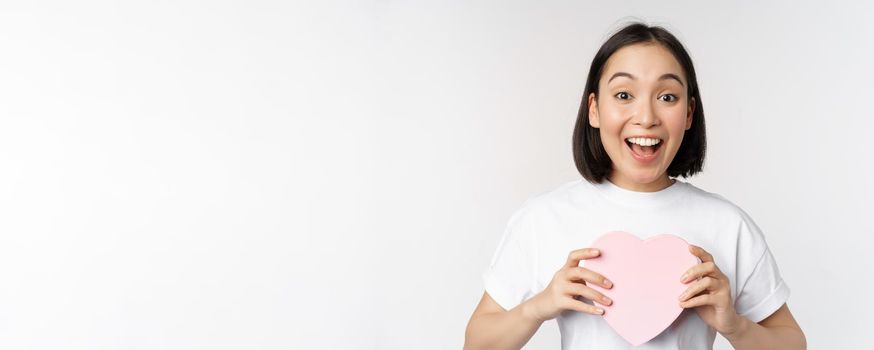 Valentines day. Happy asian girl receive romantic gift, holding heart shaped box and smiling excited, standing over white background.