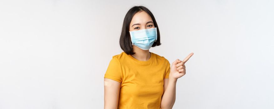 Covid-19, vaccination and healthcare concept. Portrait of cute asian girl in medical mask, has band aid on shoulder after coronavirus vaccine, standing over white background.