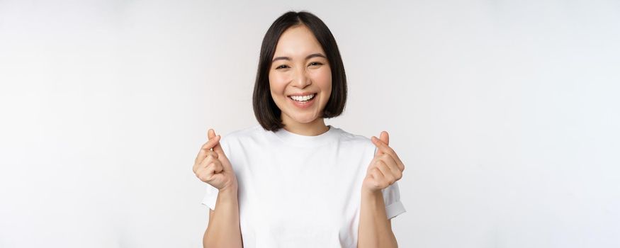 Beautiful asian woman smiling, showing finger hearts gesture, wearing tshirt, standing against white background.