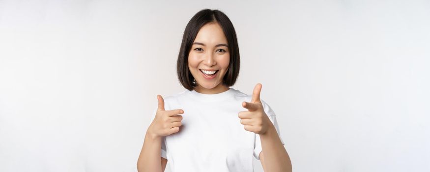 Image of smiling asian girl pointing fingers at camera, choosing, inviting you, congratulating, standing in tshirt over white background.