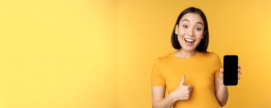 Excited asian woman showing mobile phone screen, thumbs up, like smth good, recommending smartphone application, standing over yellow background.