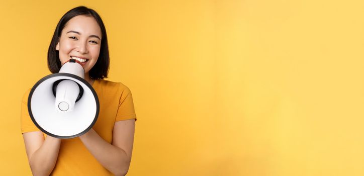 Smiling asian woman standing with megaphone, announcing smth, advertising product, standing over yellow background.