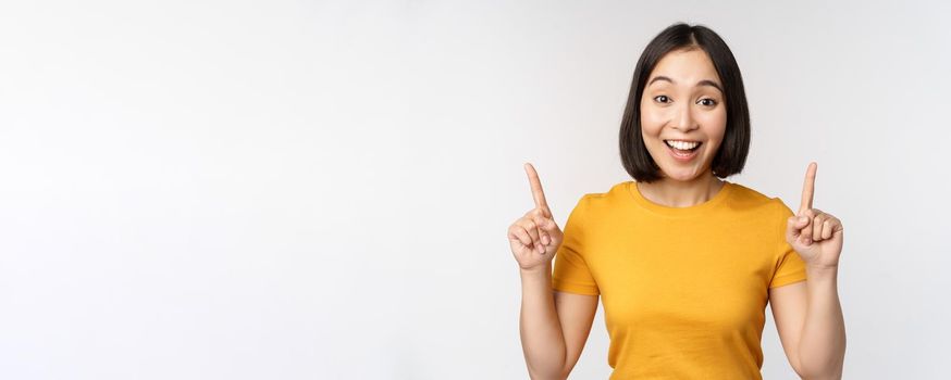Happy beautiful asian girl showing advertisement, pointing fingers up, standing over white background.