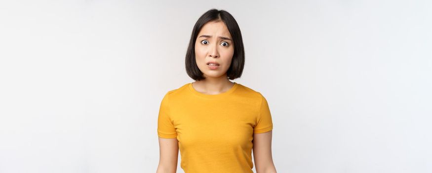 Portrait of worried korean girl, looking concerned at camera, standing in yellow tshirt over white background.