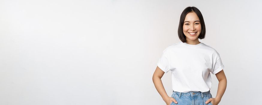 Lifestyle. Happy modern asian girl, smiling and looking happy at camera, posing in white tshirt and jeans, studio background.