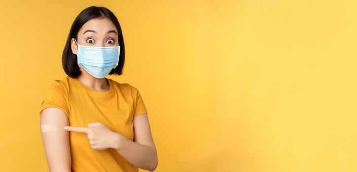 Vaccination from covid and health concept. Excited asian woman in medical face mask, pointing finger at shoulder with band aid, standing over yellow background.