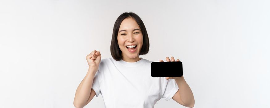 Enthusiastic asian girl scream in joy, showing horizontal smartphone screen, mobile phone display, standing over white background. Copy space