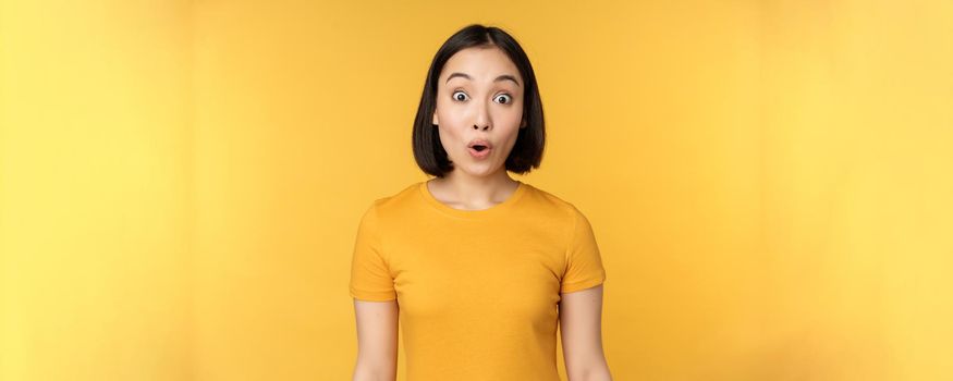 Image of asian girl looking surprised, reacting amazed, raising eyebrows impressed, standing over yellow background.
