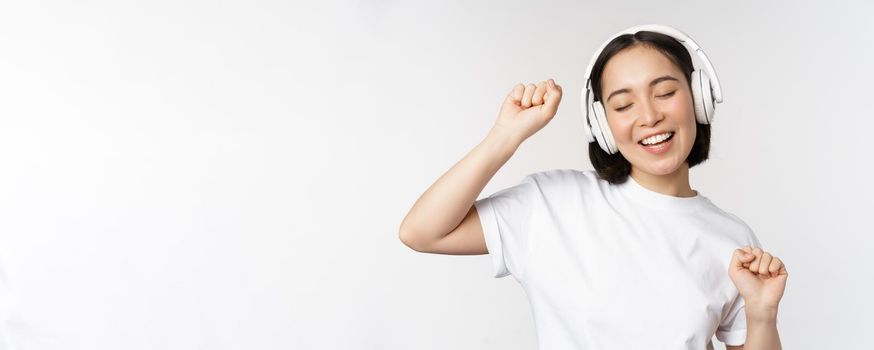 Dancing and singing asian woman, listening music in headphones, standing in earphones against white background. Copy space
