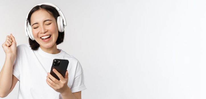 Beautiful modern asian girl, listening music in headphones, holding mobile phone, using smartphone app, standing over white background.