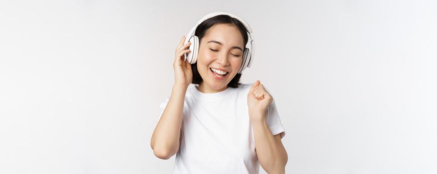 Modern asian girl dancing, listening music with headphones, smiling happy, standing in tshirt over white background.