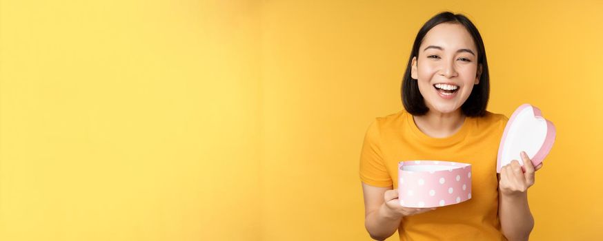 Portrait of excited asian woman, open gift box with surprised happy face, standing over yellow background.